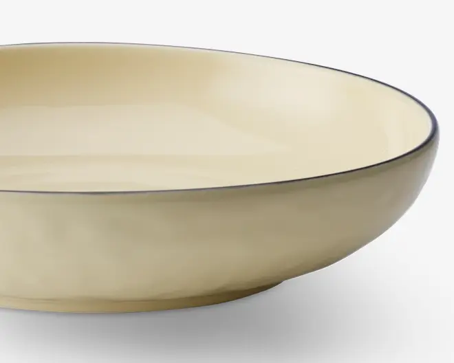 En lav, rund skål med en glat, blank cremefarvet overflade. Skålens kant er diskret markeret med en tynd, mørk streg, som giver en elegant kontrast til den ensartede farve. Skålen har blødt skrånende sider og et minimalistisk, moderne design.
