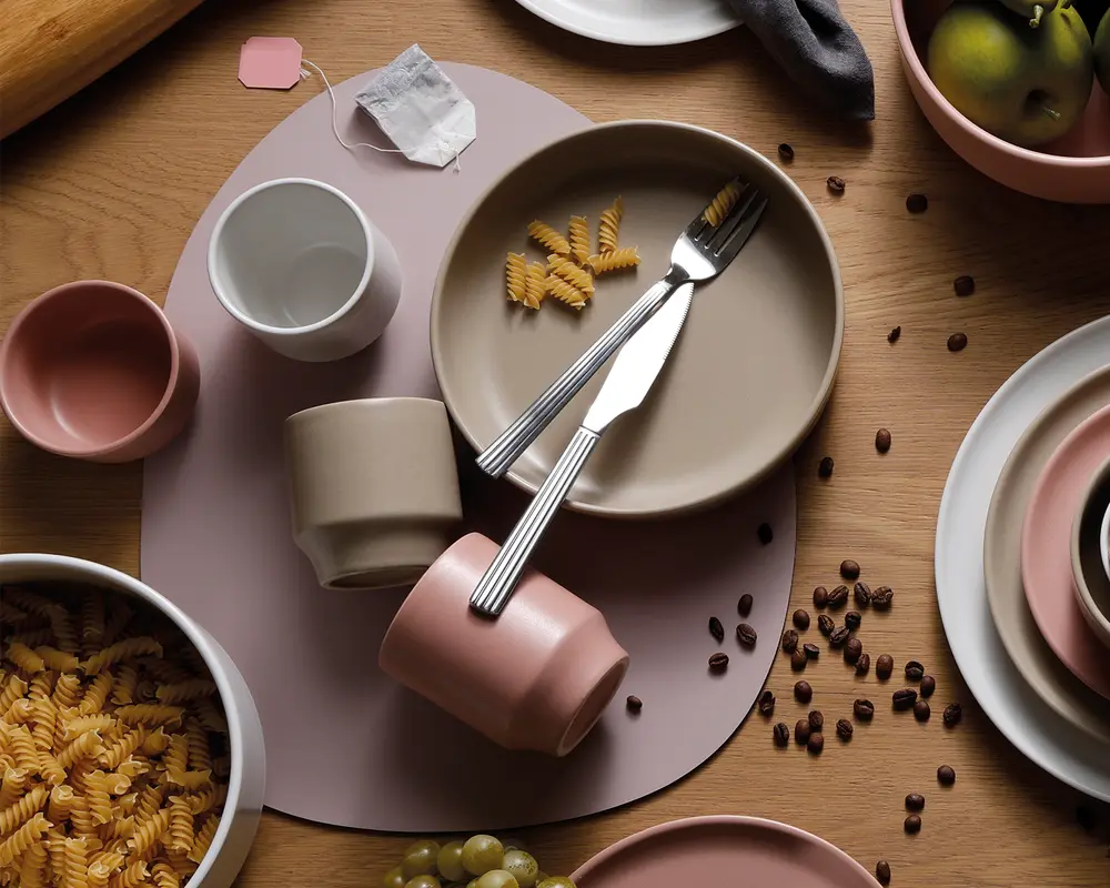 Et sæt keramikservice i afdæmpede pastelfarver, herunder tallerkener, skåle og kopper i nuancer af beige, rosa og taupe. Servicet har et minimalistisk, moderne design med glatte, matte overflader. Produkterne er arrangeret på et træbord med dækkeserviet og ledsaget af bestik i rustfrit stål. Billedet viser også ukogt pastaskruer, kaffebønner, en tepose og lidt frugt, hvilket understreger servicets alsidighed til forskellige måltider.