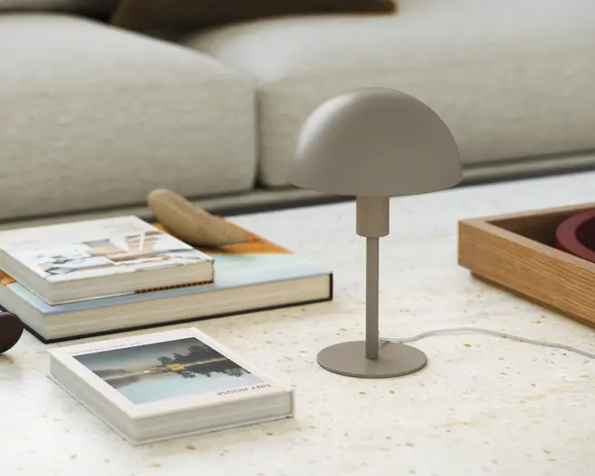 En lille, moderne bordlampe med mat taupe-finish. Lampen har en kuppelformet skærm og en slank, cylindrisk stang, der er fastgjort til en rund, flad fod. Designet er minimalistisk og nutidigt, velegnet til et skrivebord eller et sidebord. Lampen har en strømledning, der går ud fra bunden.