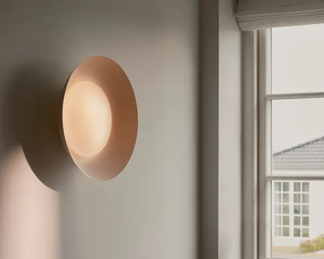 Minimalistisk væglampe med en rund yderskærm i bronzefarve og et blødt, diffust varmt lys fra midten, monteret på en neutral væg ved siden af et vindue.