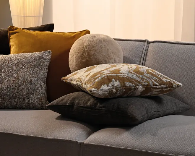 Assorterede pyntepuder i sennepsgul, grå, beige og sort med en blanding af velour, vævet stof, kunstpels og mønstrede teksturer, stablet på en lysegrå sofa.