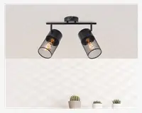 Loftmonteret lampe med to justerbare cylindriske lampeskærme, hver med et sort metalnet. Pærerne er synlige gennem nettet, hvilket giver et industrielt og moderne udtryk. Lampen er fastgjort til en rund base og en vandret stang, så lampeskærmene kan vinkles i forskellige retninger.