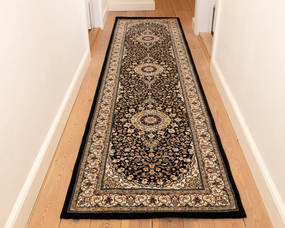 Et langt, smalt tæppe med et traditionelt persisk-inspireret design, der har en mørk bund med indviklede blomster- og vinmønstre i beige, creme og bordeaux. Tæppet har tre store, cirkulære medaljoner jævnt fordelt langs længden og en detaljeret kant med gentagne motiver. Kanterne er afsluttet med sort kantbånd, og tæppet ser ud til at have en lav luv, der egner sig til områder med meget trafik, såsom gange.