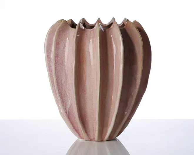 En dekorativ vase med et unikt, skulpturelt design med lodrette riller og en bølget, takket kant foroven. Vasen har en blank, lyserød-lilla glasur med diskrete farvevariationer, hvilket giver et håndlavet og kunstnerisk udtryk. Formen er rund og let tilspidset i bunden.