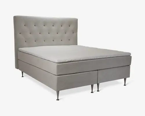 Polstret seng med høj, blød sengegavl, der har et diamantformet knapmønster. Sengen er betrukket med et lyst beige stof, har rene, lige linjer og står på slanke, metalliske ben. Madrassen er tyk og komfortabel med en ekstra topmadras for øget komfort. Udtrykket er moderne og elegant.