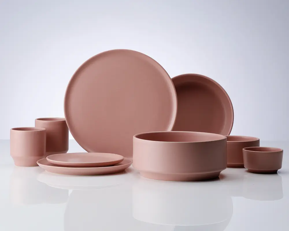 Et sæt mat, støvet rosa keramikservice med et minimalistisk design. Sættet indeholder store og små tallerkener, skåle i forskellige størrelser og kopper. Alle dele har glatte, ensfarvede overflader med blødt afrundede kanter, hvilket giver et moderne og elegant udtryk.