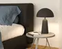 En moderne bordlampe med mat sort finish, der har en kuppelformet skærm og en cylindrisk fod. Lampen står på et rundt, lyst sidebord ved siden af en seng med mørkt polstret sengegavl. Lampens design er minimalistisk og nutidigt med rene linjer og en glat overflade.