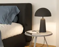 En moderne bordlampe med mat sort finish, der har en kuppelformet skærm og en cylindrisk fod. Lampen står på et rundt, lyst sidebord ved siden af en seng med mørkt polstret sengegavl. Lampens design er minimalistisk og nutidigt med rene linjer og en glat overflade.