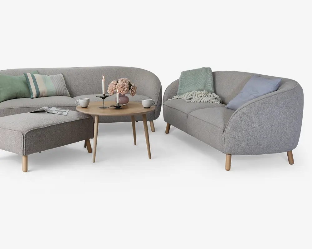 Moderne stueindretning med en lysegrå polstret hjørnesofa og en matchende to-personers sofa, begge med afrundede kanter og lyse træben. Sofaen er pyntet med puder i pastelfarver og et blødt strikket tæppe. Foran står et rundt sofabord i træ med en vase med blomster, et stearinlys, kopper og en bog, hvilket skaber en hyggelig og indbydende stemning.