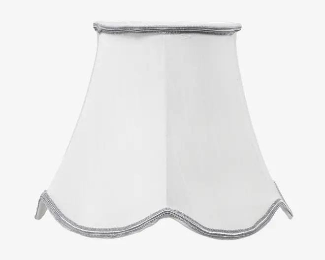 Hvid lampeskærm med en klokkeformet, udvidet design og blødt bølget kant nederst. Der er sølvfarvet kantbånd langs både top og bund, hvilket giver et diskret dekorativt præg. Stoffet er glat og let gennemsigtigt, så lyset spredes blødt.