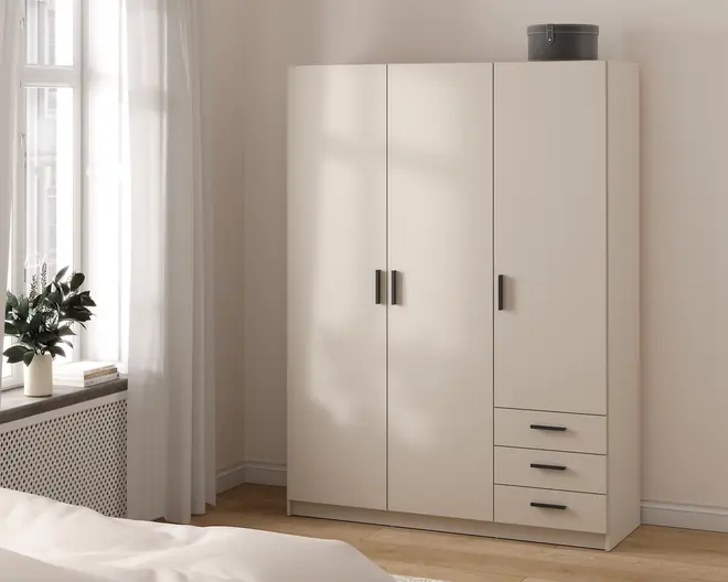 Et moderne, fritstående garderobeskab i en lys beige farve med et minimalistisk design. Skabet har tre høje låger med slanke, mørke greb og tre rummelige skuffer i højre side, alle med matchende greb. Overfladen er glat og mat, og skabet står på en flad sokkel. Øverst står en rund opbevaringsboks. Udtrykket er enkelt og moderne, velegnet til et soveværelse med neutrale farver.