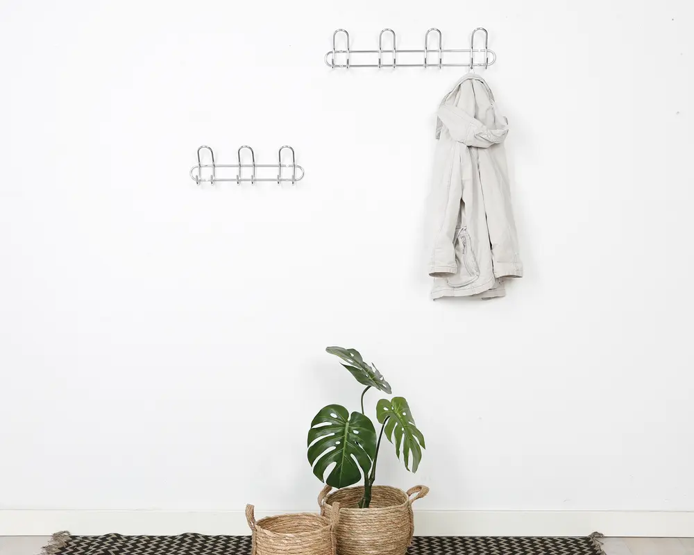 To vægmonterede metalstativer med flere kroge er fastgjort til en hvid væg. Det større stativ har fem kroge og bærer en lysefarvet badekåbe, mens det mindre stativ har tre kroge og er tomt. Nederst står to flettede kurve, hvoraf den ene indeholder en grøn monstera-plante. Gulvet har et sort og hvidt mønstret tæppe.