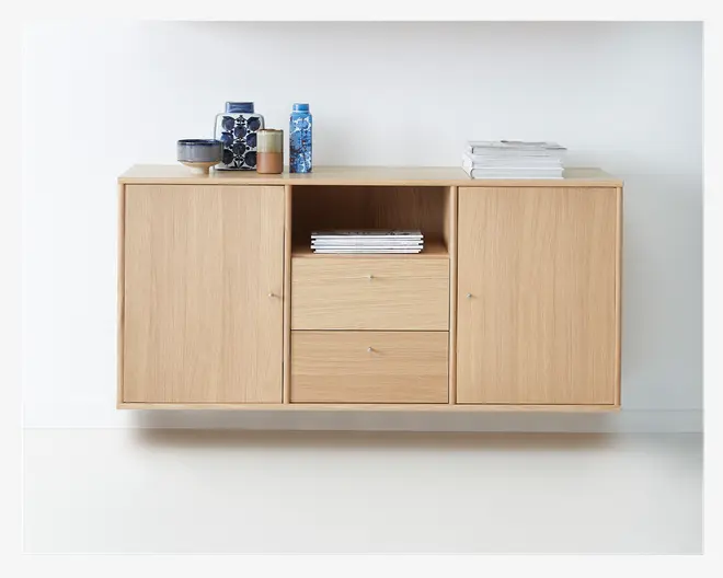 Vægmonteret skab i lyst egetræ med et minimalistisk design, der har to store låger i siderne, to centrale skuffer og en åben hylde over skufferne. Skabet har en glat, naturlig træfinish og små, runde metalgreb på låger og skuffer. Øverst på skabet står der stakke af bøger og keramiske vaser i forskellige farver og mønstre.