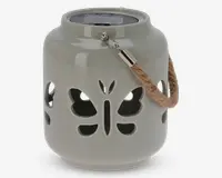 En rund, lysegrå lanterne af keramik med udskårne sommerfugleformer på siderne, som lader lyset skinne igennem. Lanternen har et kraftigt, flettet rebhåndtag fastgjort med metalbeslag, så den nemt kan bæres eller hænges op. Toppen har et forsænket område, sandsynligvis til et stearinlys eller LED-lys.