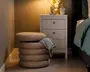 En rund, beige puf med blødt, tekstureret stof og et stablet, lagdelt design, placeret ved siden af et hvidt natbord med tre skuffer. En grå strikket plaid er lagt over puffen. Natbordet har en hvid kop, en dekorativ skulptur og en vase med hvide blomster, alt sammen oplyst af en varm lampe.