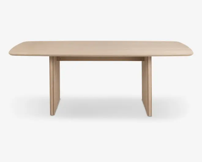 Rektangulært bord med lys træfinish, blødt afrundede hjørner og tykke, flade panelben i hver ende. Designet er minimalistisk og moderne med en glat bordplade og synlig træstruktur.
