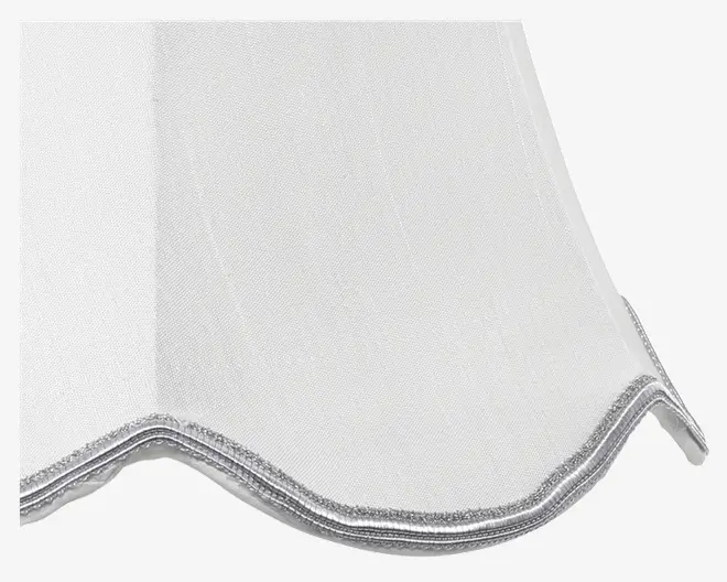 Nærbillede af et hvidt stof med let glans, der har en bølget, scallop-formet kant afsluttet med en sølvfarvet metallisk kant. Syningen langs kanten er pæn og dekorativ, hvilket giver et elegant udtryk.