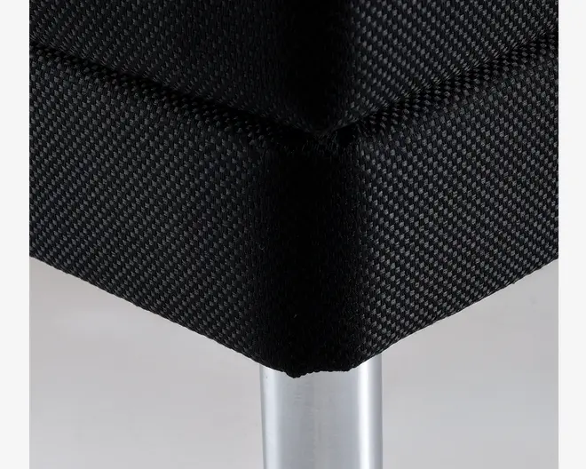 Nærbillede af et møbelhjørne med tæt vævet sort stofbetræk, der har en diskret tekstur. Hjørnet er veldefineret, og syningerne er pæne. Møblet understøttes af et cylindrisk, sølvfarvet metalben med en glat overflade.
