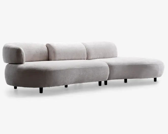 Moderne sofa med lav profil og blødt, lysebeige stofbetræk. Designet har afrundede kanter og tykke, polstrede hynder på både sæde og ryg. Venstre side har en bred, buet armlæn, mens højre side er åben, hvilket giver en chaiselong-lignende effekt. Sofaen står på korte, diskrete sorte ben, hvilket giver et svævende udtryk.