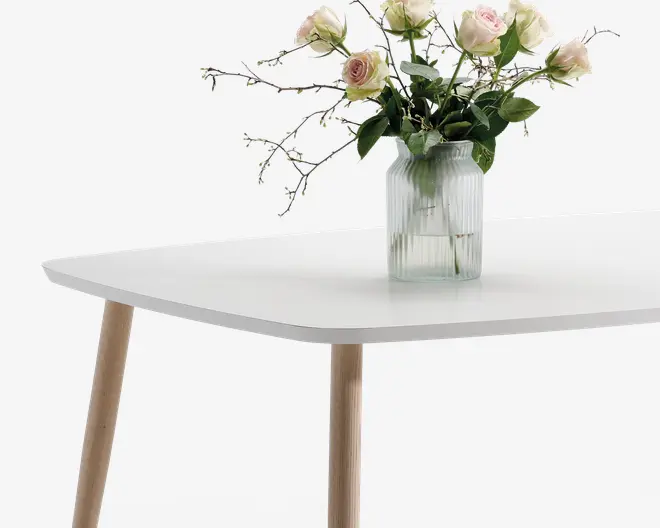 Billedet viser et bord med en glat, mat hvid rektangulær bordplade og afrundede hjørner. Bordet har skrå, lyse træben, der giver det et moderne, skandinavisk udtryk. Ovenpå bordet står en riflet glasvase med en buket lyserøde og cremefarvede roser samt grønne blade og stilke.