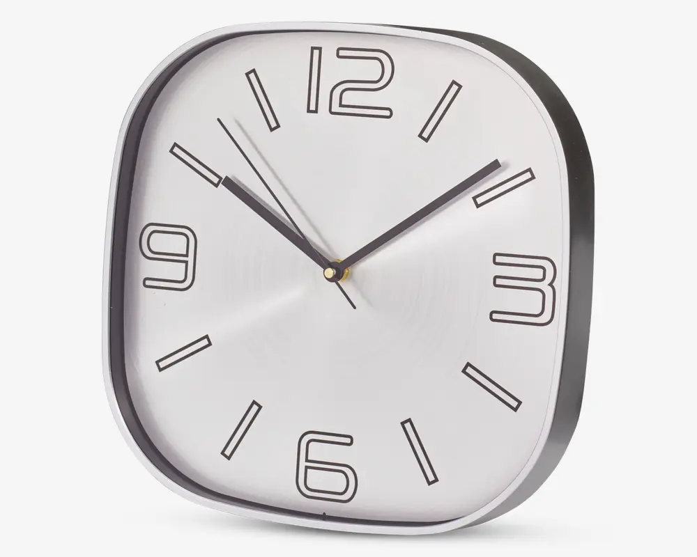 Et moderne vægur med afrundet firkantet form og sølvfarvet ramme. Urskiven er hvid med store, stiliserede sorte tal ved 12, 3, 6 og 9, samt enkle sorte streger ved de øvrige timer. Uret har tre visere: en sort timeviser, en sort minutviser og en tynd sort sekundviser. Designet er minimalistisk og let at aflæse.