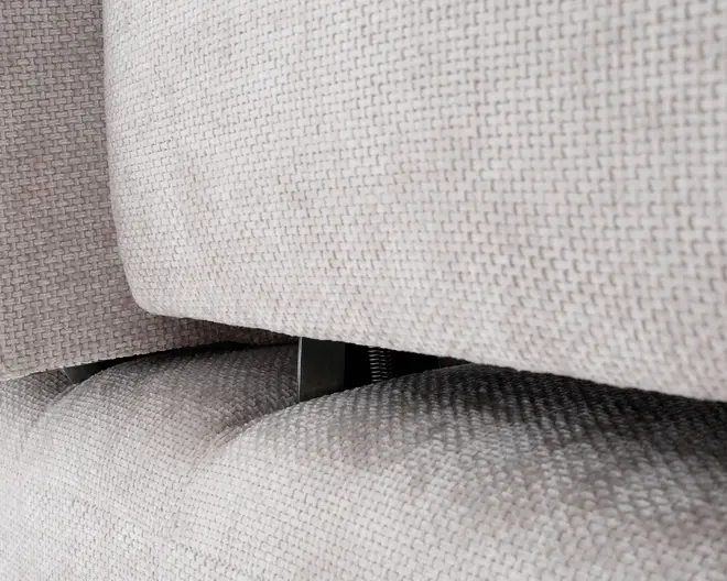 Nærbillede af en lys beige, tekstureret stofpolstring med et synligt mellemrum mellem to polstrede sektioner. Stoffet har et vævet mønster, og der ses en metalhængsel eller støtteanordning i mellemrummet, hvilket indikerer en justerbar eller tilbagelænet funktion. Hynderne fremstår tykke og bløde, hvilket antyder komfort og støtte.