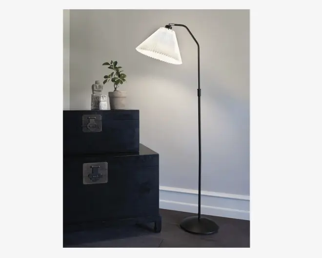Høj gulvlampe med en slank, sort metalstang og en hvid, plisseret lampeskærm, der er vinklet nedad. Lampen har en rund, stabil fod og et minimalistisk design, der passer til moderne indretning.