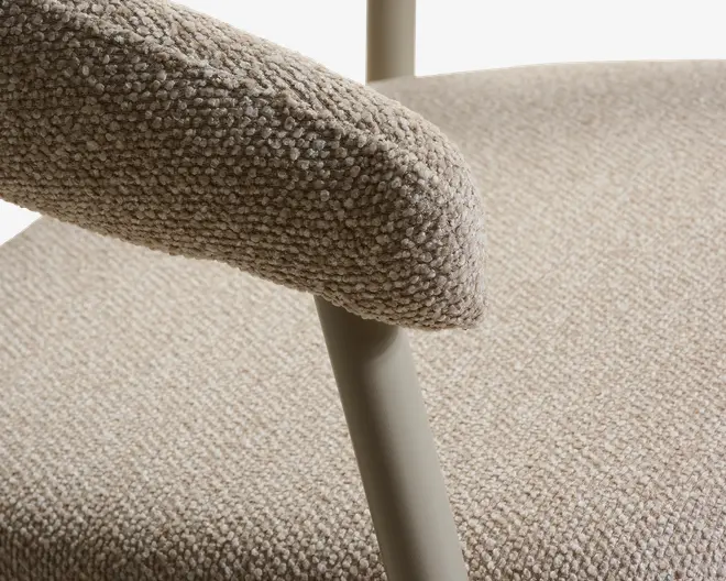 Nærbillede af et sæde betrukket med beige boucle-stof med afrundet polstret armlæn og et slankt, lys beige metalben.