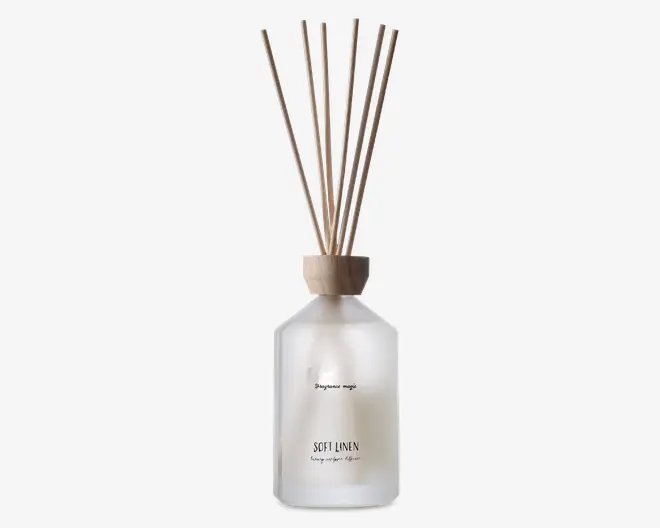 Frostet glasflaske med trælåg, der indeholder flere naturfarvede duftpinde. Flasken har minimalistisk sort tekst på forsiden, herunder ordene 'SOFT LINEN.' Det samlede design er enkelt og elegant, velegnet til moderne boligindretning.