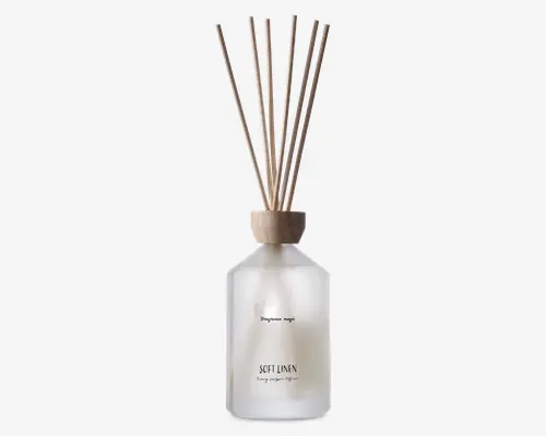 Frostet glasflaske med trælåg, der indeholder flere naturfarvede duftpinde. Flasken har minimalistisk sort tekst på forsiden, herunder ordene 'SOFT LINEN.' Det samlede design er enkelt og elegant, velegnet til moderne boligindretning.