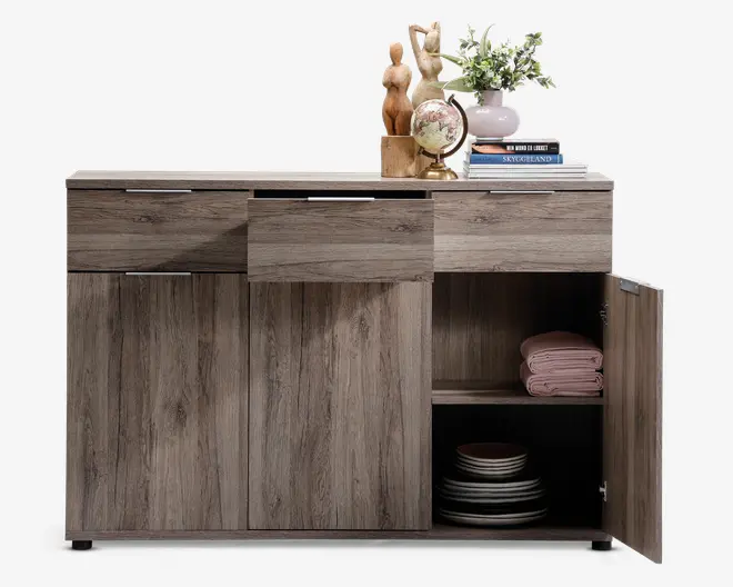 Et opbevaringsmøbel i træ med en naturlig, lysebrun finish og diskret træstruktur. Møblet har tre skuffer øverst, hvoraf den midterste er delvist åben, samt tre skabslåger nedenunder. Den højre skabslåge står åben og afslører to hylder: den øverste med pænt sammenfoldede lyserøde håndklæder og den nederste med stakke af tallerkener og skåle. Øverst på møblet står en lille globus, to træfigurer, en vase med blomster og en stak bøger.