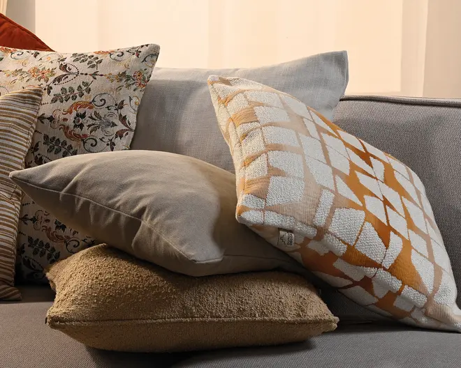 En stak dekorative pyntepuder på en grå sofa, herunder en pude med blomsterprint, en ensfarvet taupe pude, en struktureret beige pude og en creme- og orange geometrisk pude foran.