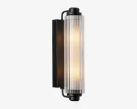Væghængt lampe med en cylindrisk, lodret glasafskærmning med rillet tekstur, der giver et diffust lys. Lampen har en mat sort metalbase og endestykker, hvilket giver et moderne og industrielt udtryk. Glasafskærmningen lader det varme lys skinne igennem og skaber en blød, stemningsfuld belysning.