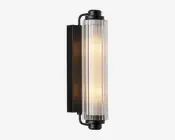 Væghængt lampe med en cylindrisk, lodret glasafskærmning med rillet tekstur, der giver et diffust lys. Lampen har en mat sort metalbase og endestykker, hvilket giver et moderne og industrielt udtryk. Glasafskærmningen lader det varme lys skinne igennem og skaber en blød, stemningsfuld belysning.