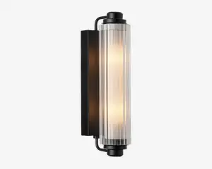 Væghængt lampe med en cylindrisk, lodret glasafskærmning med rillet tekstur, der giver et diffust lys. Lampen har en mat sort metalbase og endestykker, hvilket giver et moderne og industrielt udtryk. Glasafskærmningen lader det varme lys skinne igennem og skaber en blød, stemningsfuld belysning.