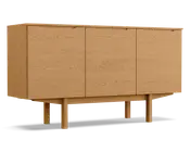 Rektangulært skab i træ med naturlig egetræsfinish, udstyret med tre flade låger med diskrete, integrerede greb øverst. Skabet står på fire solide, firkantede ben, der er forbundet af en vandret støttebjælke, hvilket giver et hævet og stabilt udtryk. Træets årer er synlige på alle flader og tilfører struktur og varme til det minimalistiske design.