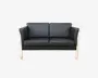 To-personers sofa med sorte læderhynder og en lys træramme. Sofaen har lige armlæn og ryglæn samt tykke, aftagelige sæde- og ryghynder for ekstra komfort. Designet er moderne og minimalistisk, hvor de synlige træben og rammen skaber en kontrast til det mørke betræk.