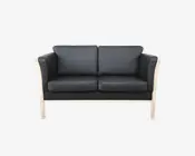To-personers sofa med sorte læderhynder og en lys træramme. Sofaen har lige armlæn og ryglæn samt tykke, aftagelige sæde- og ryghynder for ekstra komfort. Designet er moderne og minimalistisk, hvor de synlige træben og rammen skaber en kontrast til det mørke betræk.