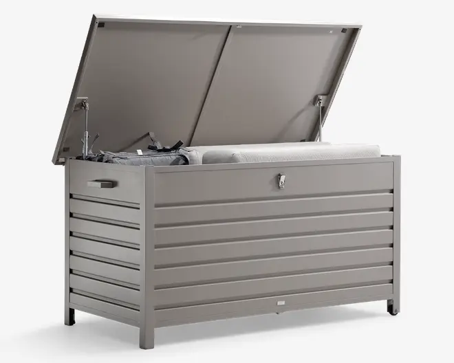 Rektangulær udendørs opbevaringsboks med hængslet låg, vist åben for at vise det rummelige indre. Fremstillet af holdbart, vejrbestandigt materiale i lysegrå farve med horisontale lameller for et moderne udseende. Har integrerede sidehåndtag for nem flytning og en låsbar haspe på fronten for sikkerhed. Indvendigt ses hynder og andet udendørstilbehør, hvilket viser den store opbevaringskapacitet.