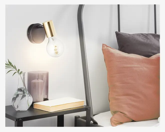 En moderne vægmonteret lampe med en mat sort, rund base og en fatning i guldfarvet metal, der holder en stor, klar, rund pære. Lampen er placeret over et sengebord, som har en glasvase med en grøn bladstilk, et lilla stearinlys og en lukket bog med cremefarvet omslag og guldkant. Sengen har en sort metalramme og er pyntet med en stor, tekstureret ferskenfarvet pude og en mindre, mørkegrå pude.