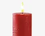 En cylindrisk, dybrød stearinlys med en tekstureret overflade og en glat, flad top. Lyset er tændt med en synlig sort væge og en klar gul-orange flamme. Voksen har en let rustik, ujævn finish, hvilket giver det et håndlavet udseende.