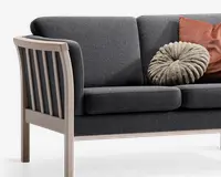 Billedet viser et close-up af en moderne sofa med mørkegrå polstrede hynder og en lys træramme. Armlænet og sidepanelet har lodrette trælameller. To pyntepuder ligger på sofaen: den ene er rund og plisseret i et blødt, lysegråt stof, og den anden er rektangulær med en rustfarvet, ribbet tekstur.