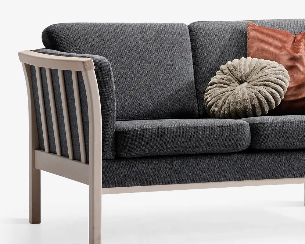 Billedet viser et close-up af en moderne sofa med mørkegrå polstrede hynder og en lys træramme. Armlænet og sidepanelet har lodrette trælameller. To pyntepuder ligger på sofaen: den ene er rund og plisseret i et blødt, lysegråt stof, og den anden er rektangulær med en rustfarvet, ribbet tekstur.