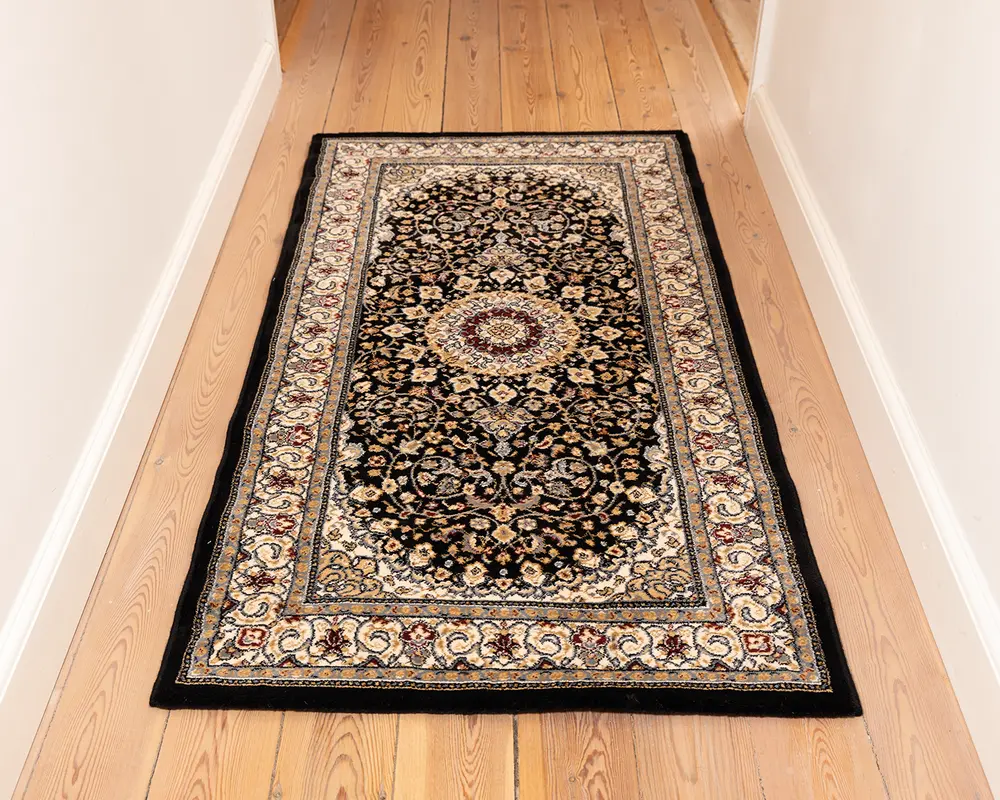 Rektangulært tæppe med et traditionelt persisk-inspireret design, der har en central medaljon i bordeaux og cremefarver omgivet af indviklede blomstermønstre i beige, guld og blå på en sort baggrund. Tæppet har en bred dekorativ kant med gentagne blomstermotiver og en sort yderkant. Teksturen ser blød og tæt ud, velegnet til områder med meget trafik som gange.