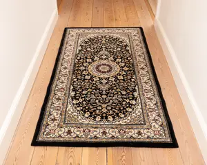 Rektangulært tæppe med et traditionelt persisk-inspireret design, der har en central medaljon i bordeaux og cremefarver omgivet af indviklede blomstermønstre i beige, guld og blå på en sort baggrund. Tæppet har en bred dekorativ kant med gentagne blomstermotiver og en sort yderkant. Teksturen ser blød og tæt ud, velegnet til områder med meget trafik som gange.