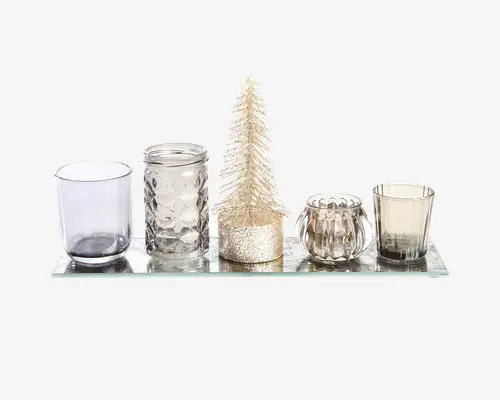 Et dekorativt sæt bestående af fem fyrfadsstager i glas med forskellige former og teksturer, arrangeret på et rektangulært spejlbakke. Fyrfadsstagerne har glatte, facetslebne og riflede designs i klare, grå og røgfarvede nuancer. I midten står et lille, glitrende guldfarvet juletræ af flaskerenser, som giver et festligt præg til arrangementet.