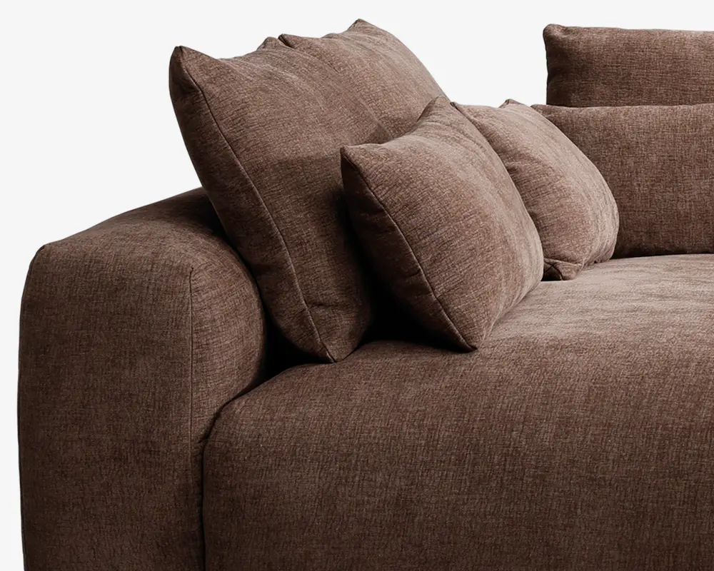 Nærbillede af et sofahjørne betrukket med tekstureret brunt stof. Sofaen har brede, bløde armlæn og leveres med flere store, fyldige ryg- og sidepuder for ekstra komfort og støtte. Stoffet har et blødt, vævet udseende, og designet er moderne og indbydende.