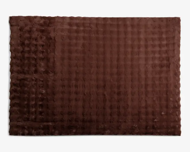 Rektangulært brunt tæppe med en blød, plysset tekstur og et diskret ternet mønster. Overfladen har hævede, tuftede sektioner langs den ene kant, hvilket giver ekstra dimension og komfort. Tæppet er ensfarvet og designet til en hyggelig, indbydende fornemmelse.