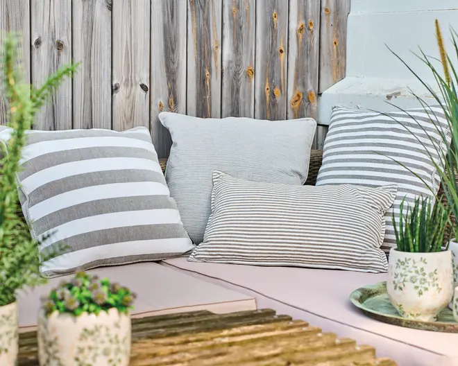 En samling pyntepuder i forskellige størrelser og former, alle i neutrale grå og hvide nuancer. Pudebetrækkene har forskellige stribede mønstre, hvor nogle har brede horisontale striber, mens andre har smalle, tætte striber. Stoffet ser blødt ud og egner sig til både udendørs og indendørs brug. Omgivelserne inkluderer en trævæg og flere grønne planter i potter, hvilket skaber en hyggelig og indbydende stemning.