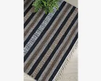 Rektangulært vævet tekstil med skiftevis sorte, brune og hvide striber samt geometriske diamantmønstre. Materialet har en tekstureret overflade, og kanterne er afsluttet med korte, cremefarvede frynser. Designet er symmetrisk, og farverne er jordnære, hvilket passer godt til en rustik eller bohemeagtig indretning.